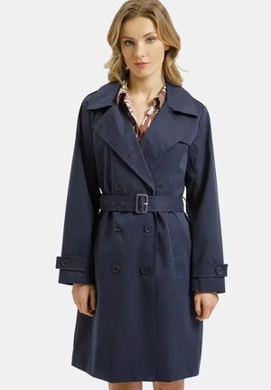 Kobieta ubrana w granatowy dwurzędowy płaszcz trencz z paskiem, szerokimi klapami i mankietami na guziki, na wzorzystej koszuli.