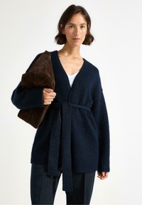 Cardigan lavorato a maglia blu navy con scollatura a V profonda e vita cin cin, caratterizzato da una texture morbida e maniche lunghe, abbinato a pantaloni a righe.