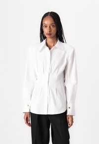 Camisa blanca ajustada de manga larga, con cuello puntiagudo y costuras decorativas. Presenta acentos de botones dorados y un diseño de frente plisado.