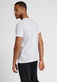 Camiseta de algodón blanca con cuello redondo, mangas cortas y un corte relajado, mostrada desde atrás sobre un fondo neutro.