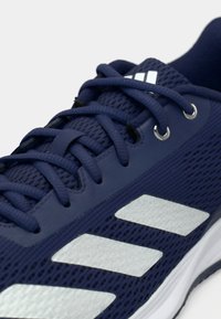 Gros plan sur une chaussure de sport bleu marine en tissu mesh, avec des lacets assortis, trois bandes blanches inclinées et des œillets métalliques.