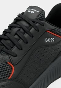 Black sneaker made of textured synthetic leather with mesh detailing, red accent, tonal laces, and branding on the tongue and side.