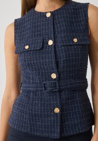 Vesta din tweed bleu marin cu un model texturat, având un gât înalt, butoane aurii și două buzunare frontale. Include o talie cu curea.