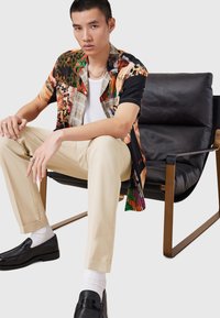 Camisa de manga corta, multicolor en negro, naranja y verde, combinada con pantalones beige y mocasines negros, sentado en una silla moderna.