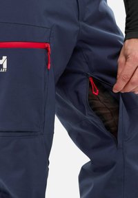 Pantalons imperméables de couleur marine avec des zips rouges, dotés d'une poche avant et d'un zip de ventilation sur le côté, fabriqués en tissu durable et texturé.