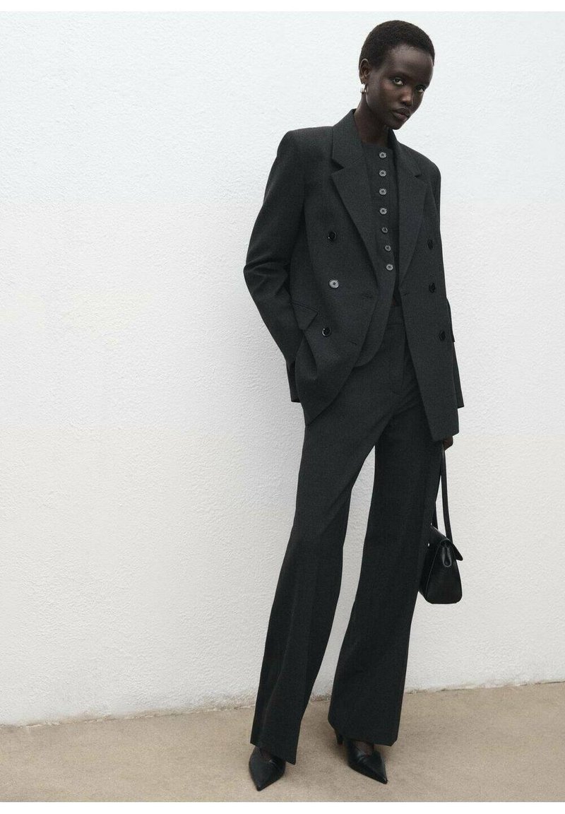 Blazer negro ajustado de dos filas de botones y pantalones de pierna ancha hechos de tela texturizada; combinado con una blusa ajustada de botones y tacones negros puntiagudos.