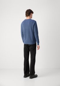 Blå genser med rund hals, teksturert stoff og ribbede mansjetter, kombinert med svarte bukser og mørke sko, sett fra baksiden.