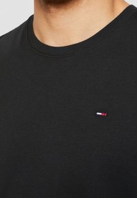 Detailní záběr na osobu, která má na sobě černé tričko s kulatým výstřihem, na kterém je na hrudi malé obdélníkové logo v červené, bílé a modré barvě.