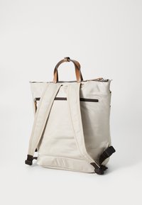 Sac à dos en tissu beige avec des accents en cuir marron, doté de bretelles ajustables, d'une poignée supérieure et d'une poche zippée à l'arrière pour le rangement.
