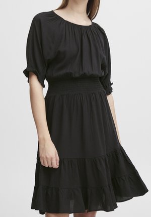 Frau in einem schwarzen Kleid mit geraffter Taille, kurzen, aufgeplusterten Ärmeln und einem gestuften, voluminösen Rock vor einfarbigem Hintergrund.