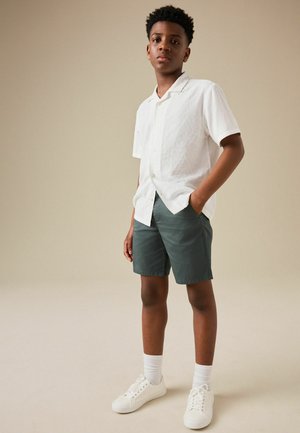 Teen boy standing with one hand in pocket, wearing white short-sleeve button-up shirt, dark shorts, white socks, and white sneakers.