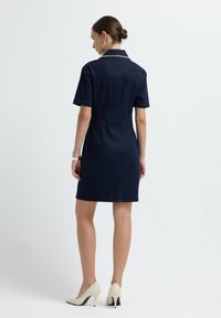 Granatowa sukienka dżinsowa z krótkimi rękawami i kołnierzykiem, z teksturowanym akcentem przy dekolcie. Noszona z jasnymi szpilkami.