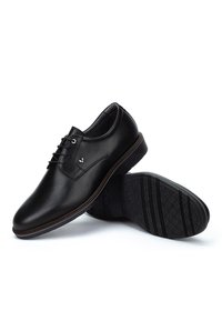 Scarpe eleganti in pelle nera con lacci e finitura liscia, una scarpa eretta e l'altra che mostra la suola texturizzata su uno sfondo bianco.