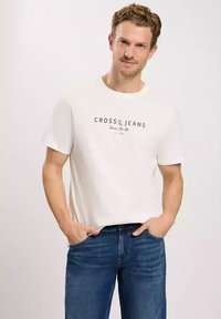 Biała bawełniana koszulka z logo "CROSS JEANS" w kolorze czarnym, w połączeniu z niebieskimi dżinsami. Koszulka ma klasyczny okrągły dekolt i krótkie rękawy.