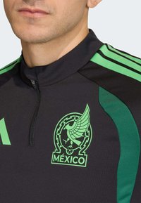Fekete sportpóló zöld díszítésekkel, cipzáros nyakkal, feltűnő zöld logóval, amely egy szárnyas alakot és a "MÉXICO" szót ábrázolja.