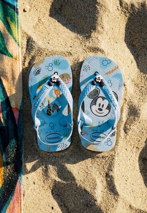 Lasten siniset Havaianas-sandaalit, joissa on sarjakuvahahmoja ja Mikki Hiiri -kuvioita, asetettuna hiekkarannalle värikkään pyyhkeen viereen.