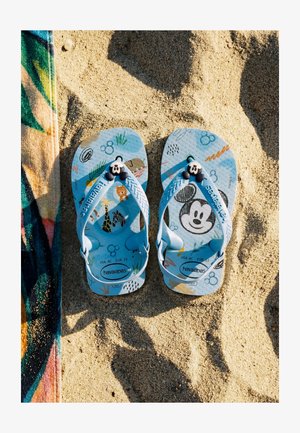 Blaue Kinder-Havaianas-Sandalen mit Cartoon-Tier- und Mickey-Mouse-Designs, auf einem Sandstrand neben einem bunten Handtuch platziert.