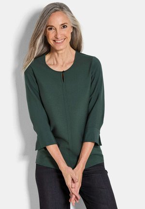 Woman with long gray hair smiles, wearing a dark green 3/4 sleeve top and dark pants, standing against a white background.