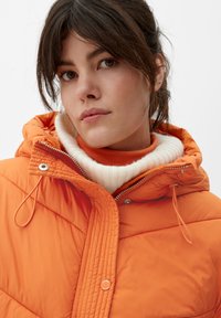 Oranje pufferjack met een aansluitende capuchon, voorzien van een ritssluiting, een geribbelde witte kraag en een gestructureerde stiksels op de borst.