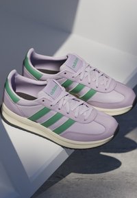 Par de zapatillas Adidas de color púrpura claro con rayas verdes y logo, medias suelas blancas y suelas exteriores negras colocadas sobre una superficie blanca inclinada.