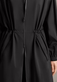 Zwarte lichtgewicht jas met een rits aan de voorkant, elastische tailleband en zijzakken met elastische manchetten voor een goede pasvorm. Matte textuur.