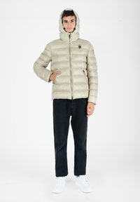 Beige pufferjakke med høj krave og hætte, med vandret quiltede sektioner og lynlås foran. Båret over sorte bukser og hvide sko.