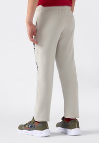 Person som har på seg beige joggebukser med sidelomme og olivengrønne joggesko med hvite såler og røde sokker, står på en hvit bakgrunn.