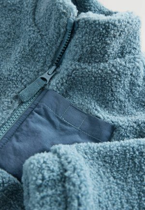 Fleecejacke in sanftem Blau mit hohem Kragen, Reißverschluss und einer kontrastierenden Tasche aus glattem Stoff, die sichtbare Nähte aufweist.