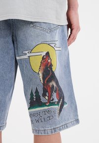 Shorts de mezclilla con un gráfico de un lobo aullando contra una luna amarilla y árboles, con el texto "NACIDO PARA SER SALVAJE" en negro.