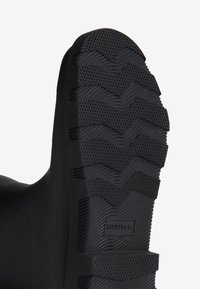 Semelle en caoutchouc noir avec un motif texturé, des rainures profondes et des accents surélevés. Logo de la marque "HUNTER" visible sur le côté.