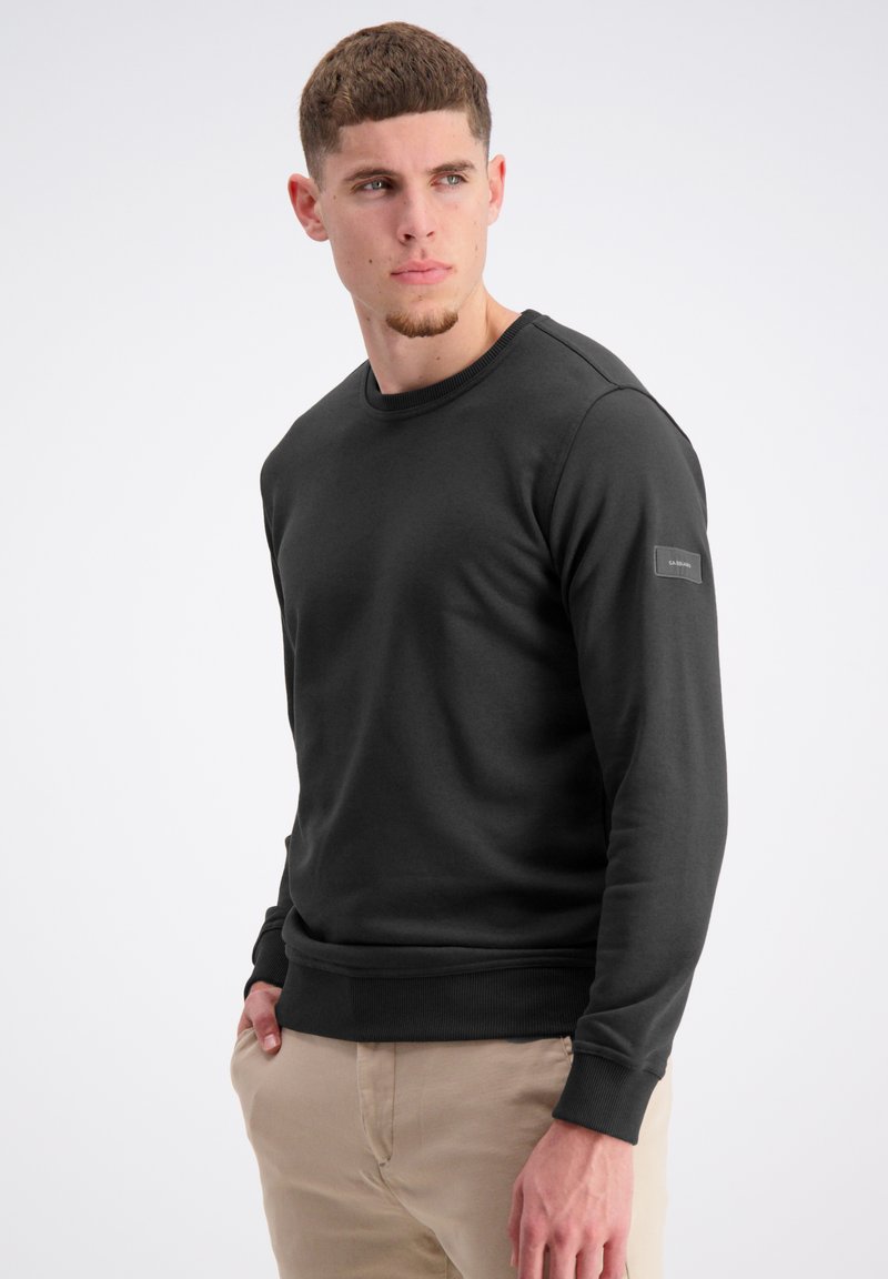 Mörkgrå sweatshirt med rund halsringning, ribbade muddar och nederkant samt en textilflik på vänster ärm, tillverkad av mjukt material.