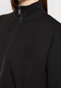 Zwarte zip-up jack met een hoge kraag, gemaakt van een zachte stof. Kenmerkend is een subtiel logo en een strakke ritssluiting.