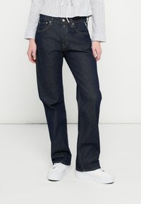 Temno modre jeans hlače iz jeansa s ravnim krojem hlačnic, z sprednjimi žepi in klasičnim zapenjanjem na gumbe, nošene z belimi supergami.