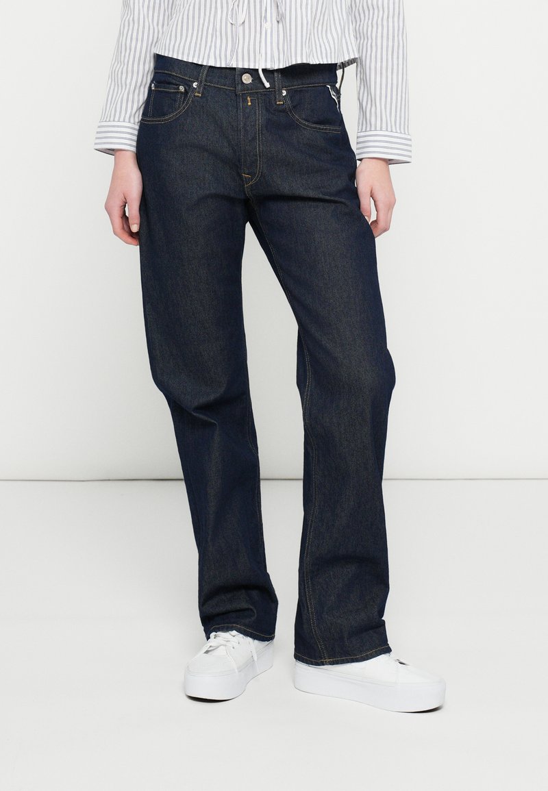 Jeans di denim blu scuro con taglio a gamba dritta, dotati di tasche anteriori e una classica chiusura con bottoni, indossati con sneakers bianche.