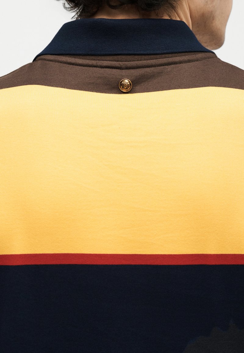 Back view of a person wearing a color-blocked shirt with navy collar, brown, yellow, red, and blue horizontal stripes, and a small gold button at the neck.