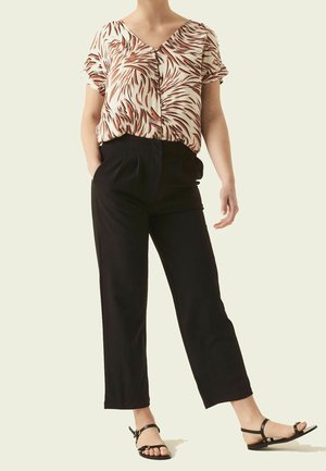 Kobieta w beżowo-brązowej bluzce z krótkim rękawem, czarnych spodniach z wysokim stanem oraz czarnych sandałach z paskami wokół kostki.