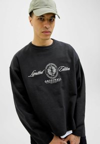 Fekete pulóver kerek nyakkal és oversized szabással. Fehér hímzett szöveggel rendelkezik, amelyen az "Limitált Kiadás" és az "ORIGINALS" felirat szerepel. Puha, texturált anyag.