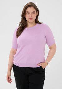 Bluza tricotată mov deschis cu mâneci scurte și decolteu rotund. Textură netedă, croială mulată, asortată cu pantaloni negri și brățară argintie.