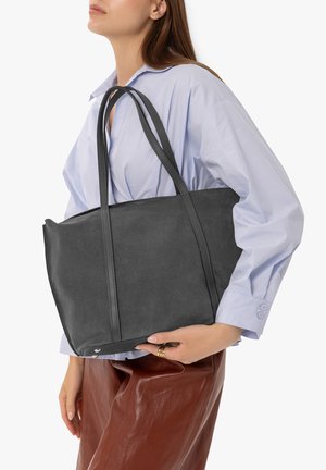 Woman wearing light blue shirt and brown leather pants holding a large black suede tote bag over her shoulder.