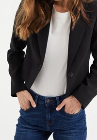 Blazer noir ajusté avec un bouton et un col, porté par-dessus un t-shirt blanc et un jean en denim foncé avec un bouton à l'avant et des détails de poches.
