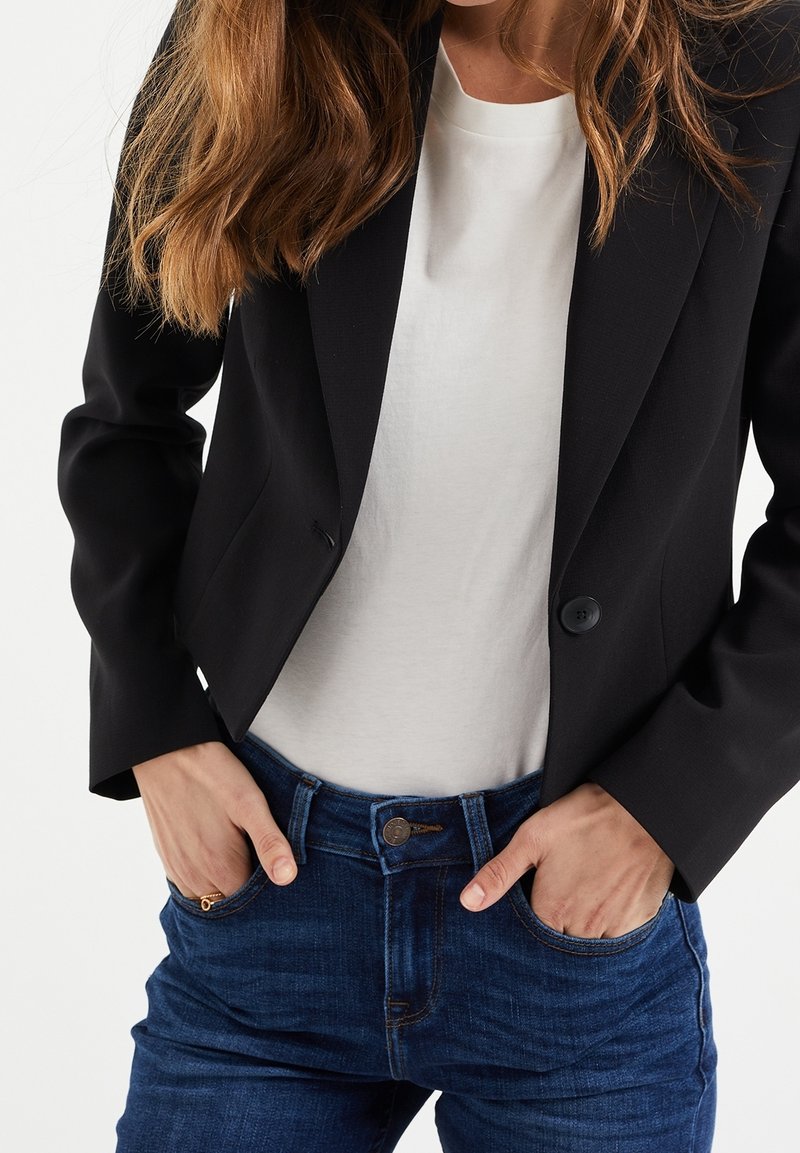 Blazer noir ajusté avec un bouton et un col, porté par-dessus un t-shirt blanc et un jean en denim foncé avec un bouton à l'avant et des détails de poches.
