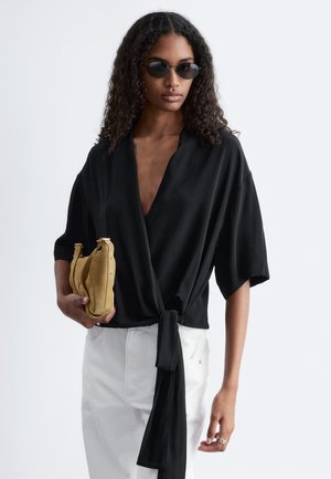 Kobieta z kręconymi włosami w czarnej bluzce kopertowej i okularach przeciwsłonecznych, trzyma beżową torebkę, ubrana w białe spodnie na jednolitym tle.