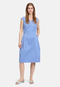 Himmelblaues, knielanges Kleid mit kurzen Ärmeln, V-Ausschnitt, seitlichen Taschen und tailliertem Schnitt, aus glattem Material. Kombiniert mit himmelblauen Sandalen.