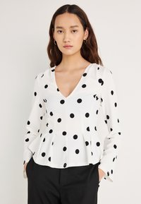 Hvit langermet bluse med svarte polkadotter, V-hals og peplumkant. Glatt stoff med en avslappet passform, typisk for fritidsbruk.