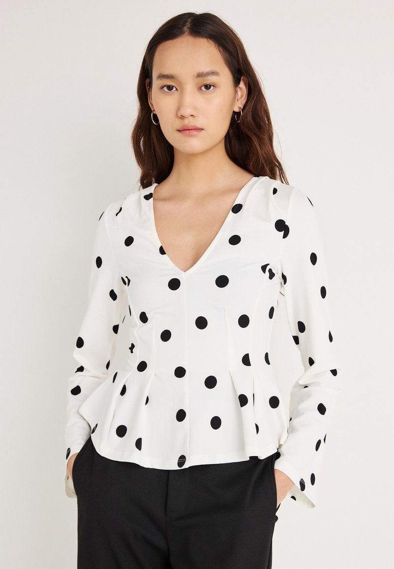 Hvit langermet bluse med svarte polkadotter, V-hals og peplumkant. Glatt stoff med en avslappet passform, typisk for fritidsbruk.