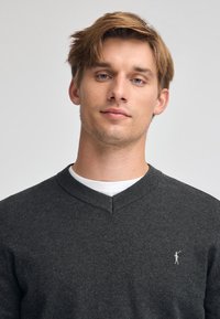 Mörkgrå v-ringad tröja tillverkad av mjukt tyg. Har ribbade ärmslut och en liten vit logotyp broderad på vänster bröst.