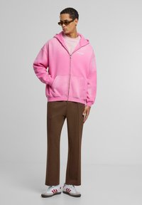 Rosa gradient zip-up hettegenser med frontlommer, sammen med brune vide bukser og hvite sneakers med røde detaljer. Modell iført solbriller.
