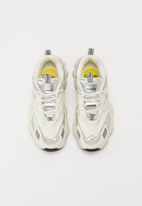 Baskets de sport blanches et argentées avec un dessus texturé, une semelle épaisse et des détails de marques prononcés. Lacets et accents de logo bicolores visibles.