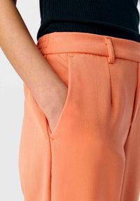 Pantalon de couleur pêche avec un tissu lisse et légèrement texturé. Comprend des poches latérales, une coupe slim et une taille avec passants de ceinture.