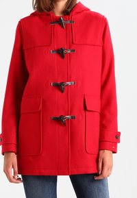 Rode wollen jas met capuchon, voorzien van sluitingen met koorden, twee voorzakken en manchetten met knopen. Gladde textuur en een getailleerde pasvorm.